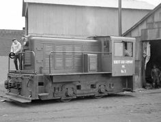 black and white photograph of a man standing on the side of a train in front of a building