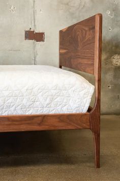 a wooden bed frame with white sheets in a concrete walled room and cement wall behind it