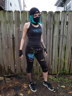 a woman with blue hair wearing a black shirt and knee length pants standing in front of a wooden fence