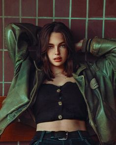 a woman in black shirt and green jacket leaning on wall with her arms behind her head