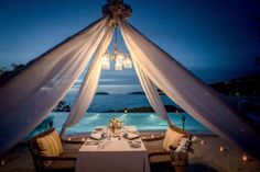 an outdoor dining area with a table and chairs under a canopy over the pool at night