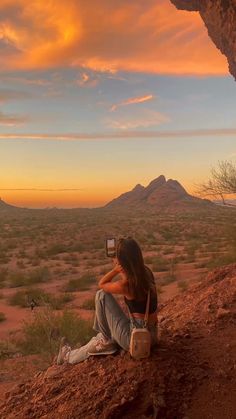 a woman sitting on the side of a cliff taking a photo with her cell phone