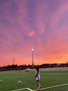 a woman standing on top of a lush green field next to a soccer ball under a pink sky