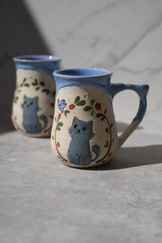 two mugs with cats painted on them sitting next to each other in front of a marble wall