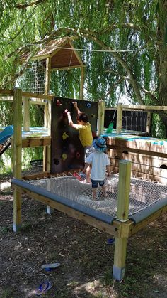 two children playing on a climbing wall in the shade under a large leafy tree