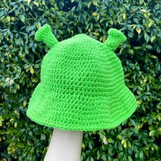 a green crocheted hat on top of a white hand with bushes in the background