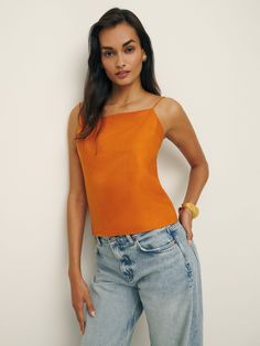 a woman standing in front of a white wall wearing jeans and an orange tank top