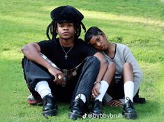 two people sitting on the grass with one person wearing a black hat and another in plaid pants