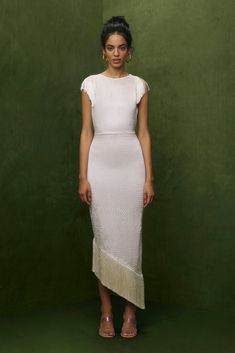 a woman standing in front of a green background wearing a white dress with fringes