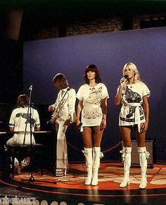 three women in short skirts and white boots on stage with microphones, one standing behind the other