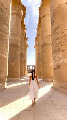 a woman in white dress walking between two large pillars at an ancient city with columns on both sides