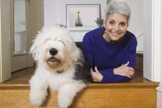 an older woman sitting next to a white dog on top of a wooden table with her arms crossed