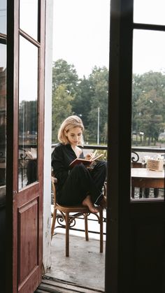 a woman is sitting in a chair reading a book on the porch outside her house