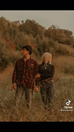 a man and woman holding hands walking through tall grass with trees in the back ground