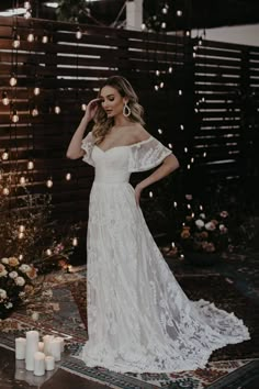 a woman standing in front of candles wearing a white wedding dress with off the shoulder sleeves