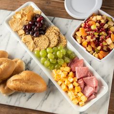 two trays filled with different types of food on a marble counter top next to bowls of fruit and bread