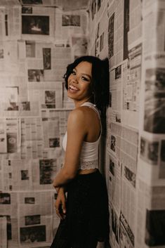 a woman standing in front of a wall covered with newsprinted papers and smiling at the camera