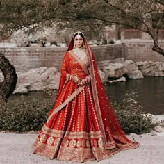 a woman in a red and gold bridal gown standing by a river with trees