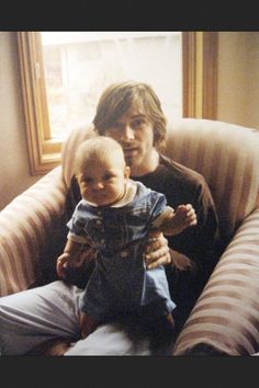 a man holding a baby in his lap while sitting on a couch next to a window