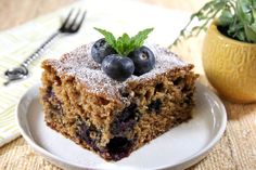 a piece of blueberry cake on a plate next to a fork and potted plant