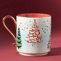 a coffee cup with christmas trees painted on it's side and gold handles, against a pink background