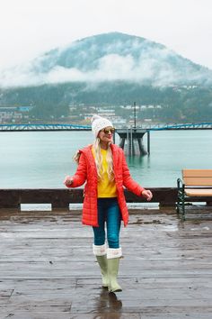 a woman in red jacket and boots standing on wooden deck next to water with mountains in the background