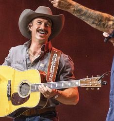 a man with a cowboy hat holding a guitar and pointing to another man in the background