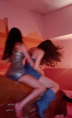 two women are dancing on a bed in a room with pink walls and orange accents