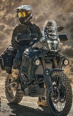 a man riding on the back of a black motorcycle down a dirt road next to a mountain