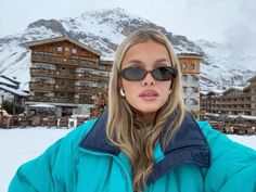 a woman wearing sunglasses standing in front of a building with snow covered mountains behind her