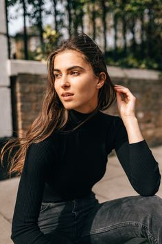 a young woman sitting on the ground wearing black jeans and a turtle neck sweater, with her hair blowing in the wind