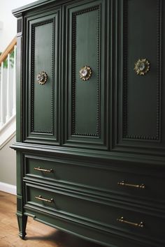 a green armoire with brass handles and knobs on the doors, in front of a staircase