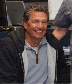 a man sitting in front of a computer desk smiling at the camera with his hands on his hips