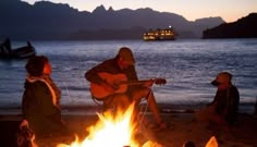 three people sitting around a campfire with a boat in the background