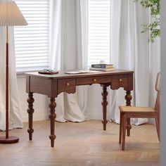 a wooden desk sitting next to a window covered in white drapes and a lamp