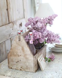 a wooden box with two bottles in it on a table next to flowers and a lamp