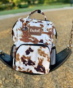 a brown and white cow print backpack sitting on a swing in a park with the name rachel written on it