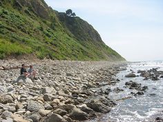 two people standing on rocks near the water and mountains with green grass growing on them