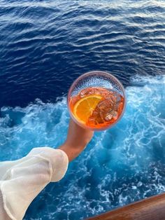 a person is holding up a glass with an orange slice in it on the deck of a boat