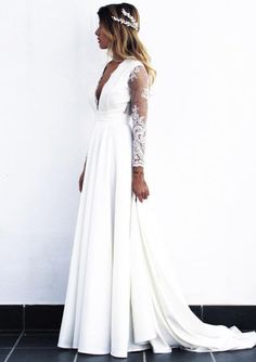 a woman standing in front of a white wall wearing a wedding dress with long sleeves