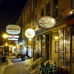 a snowy street lined with shops and businesses at night time in the winter, lit by christmas lights