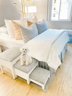 a white dog sitting on the foot stools in front of a bed with pillows