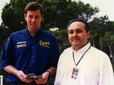 two men standing next to each other in front of trees and one is holding a camera