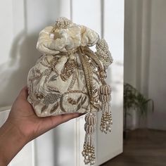 Add a touch of elegance and sophistication to your bridal look with our exquisite Cream Velvet Potli Bag. Crafted from premium velvet, this potli bag is a testament to timeless beauty and unmatched craftsmanship. The soft cream shade exudes a subtle charm that complements any bridal attire, while the intricate hand-embroidered zardozi and sequins work create a mesmerizing display of artistry. Each stitch and sequin is meticulously placed, showcasing the skill and dedication of our artisans. Designed for comfort and style, this potli bag features a stunning handmade braided handle adorned with delicate beads, offering both durability and a unique aesthetic. The handle is carefully crafted to ensure a comfortable grip, making it an ideal accessory for the bride on her special day. To further Elegant Handheld Beaded Evening Bag, Elegant Silver Embroidered Bags, Elegant Pearl White Beaded Clutch, Elegant White Shoulder Bag Gift, Handheld Pearl Embroidered Evening Bag For Weddings, Handheld Pearl Embroidery Evening Bag For Wedding, Handheld Evening Bag With Pearl Embroidery For Wedding, Elegant Pearl White Clutch With Pearl Embroidery, Cream Evening Bag With Pearl Handle For Gift