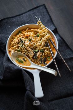 a white bowl filled with noodles and chicken on top of a black cloth next to two silver spoons