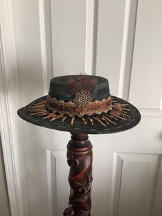 This is a custom weathered hat with incredible detail.  Black suede hat weathered in green.  Incredible fringe trim of tan beads is a show stopper.  Also has multi colored fabric trim and a feather band.  Includes feathers, vintage button and wood flower adornment.  Rim is trimmed in hemp. Adjustable band in side. Vintage Fedora Mini Hat For Festivals, Brown Vintage Fedora For Festival, Vintage Feather Hat Bands For Western-themed Events, Vintage Feather Hat Bands For Country Events, Vintage Hat Bands With Feathers For Country Events, Vintage Feather Hat Bands For Festival, Vintage Fedora With High Crown For Festivals, Vintage High Crown Fedora For Festivals, Vintage Brown Costume Hats And Headpieces For Festival