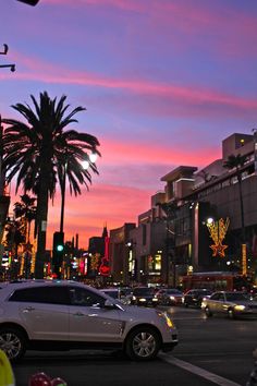 a city street filled with lots of traffic under a colorful sky at dusk, and palm trees in the foreground