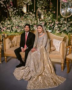 a man and woman sitting on a couch in front of a floral wall with flowers