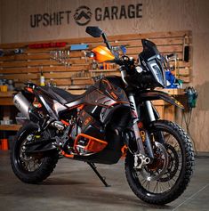 an orange and black dirt bike parked in a garage next to a wooden shelf with tools on it