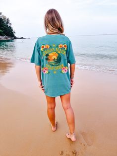 "Introducing the stunning Laguna Beach California tee! Perfect for all those who love the tropical vibes and laid-back beach lifestyle. Featuring a gorgeous coconut girl aesthetic, this shirt is adorned with an array of beautiful hibiscus, plumeria, and other tropical plants, creating a lush and vibrant design that's sure to turn heads. At the center of the design, you'll find an illustration of someone surfing at sunset, capturing the essence of the iconic California surf culture. The warm colo Tropical Graphic Print T-shirt For Beach, Cotton Tropical Print T-shirt, Cotton T-shirt With Tropical Print, Tropical Print T-shirt For Beach In Summer, Tropical Print T-shirt For Beach Season, Hawaiian T-shirt With Tropical Print For Beach, Hawaiian T-shirt For Surfing In Summer, Blue Beach T-shirt With Front Print, Tropical Print T-shirt For Beach